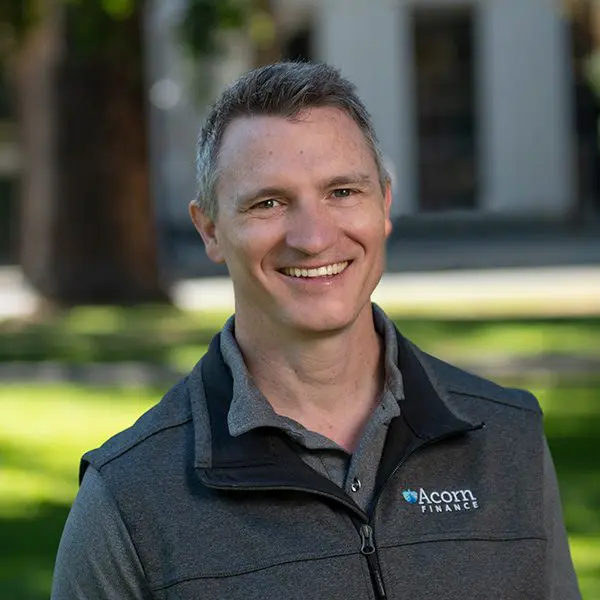 Man outdoors in jacket with "acorn finance" logo, smiling.
