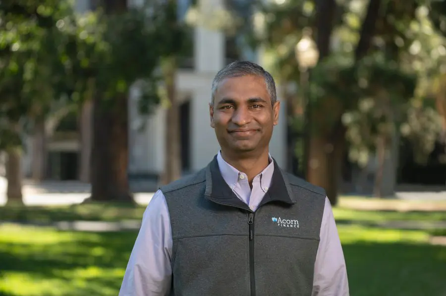 Man in Acorn Finance vest stands outdoors by building, trees.
